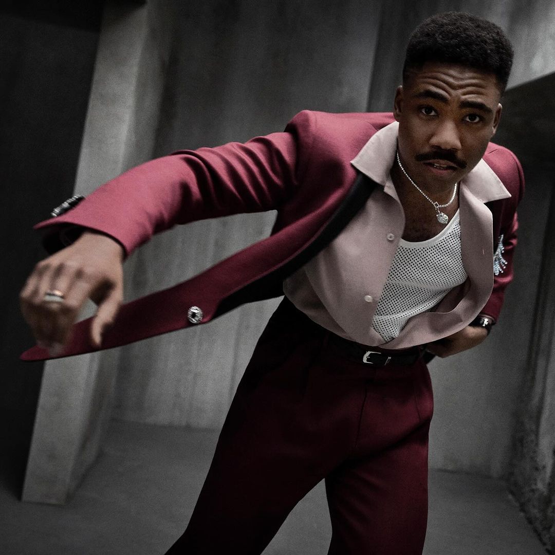 Stylish man in a maroon suit poses in a dimly lit room, showcasing fashion and style with a confident expression and eye-level framing.