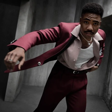 Stylish man in a maroon suit poses in a dimly lit room, showcasing fashion and style with a confident expression and eye-level framing.