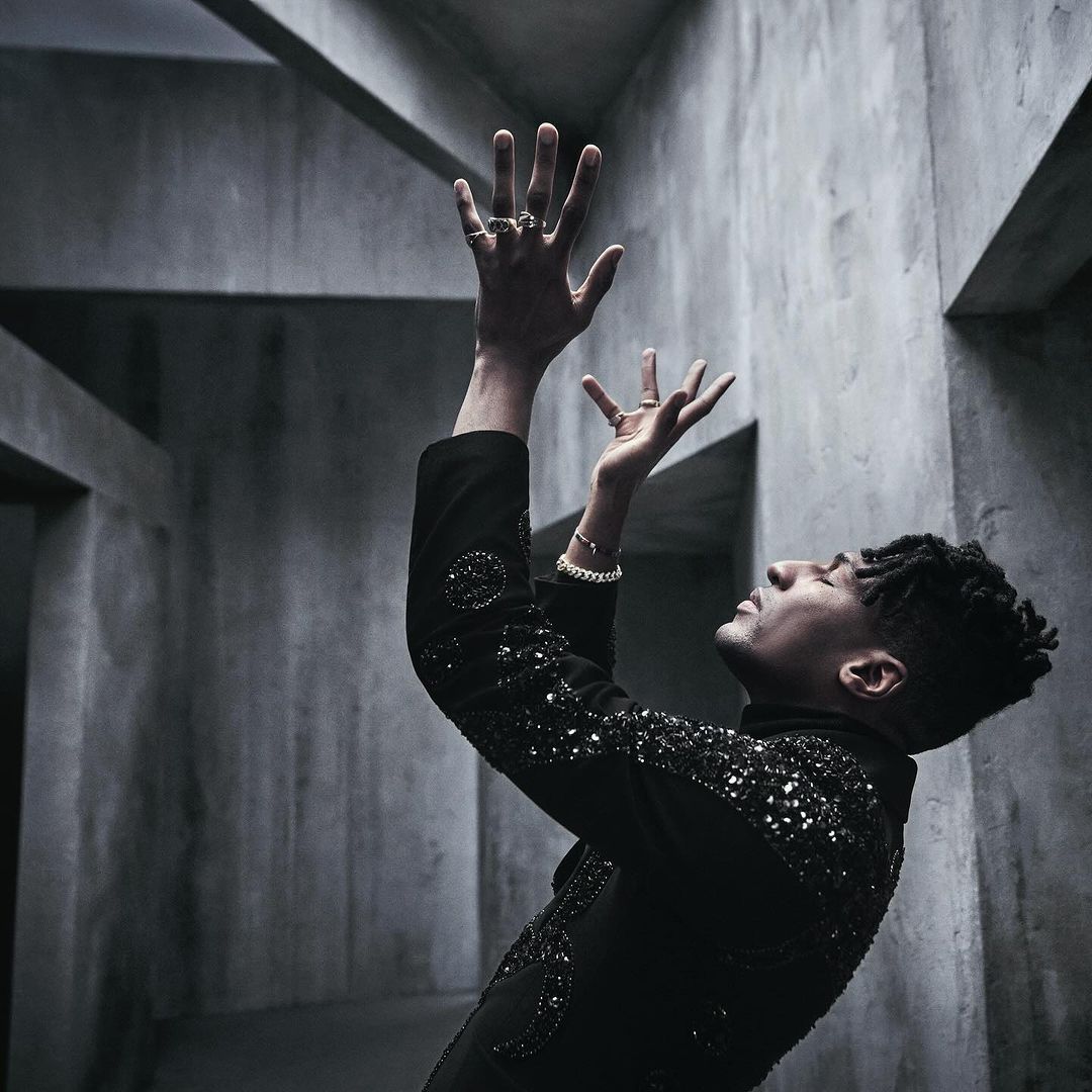 Side view of an adult male in dark embellished jacket with raised hands in a grey geometric architectural interior setting. Moody lighting.