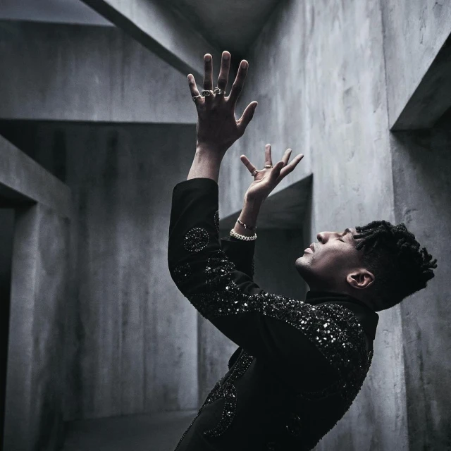 Side view of an adult male in dark embellished jacket with raised hands in a grey geometric architectural interior setting. Moody lighting.