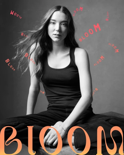 A young woman with long brown hair is seated in black clothing against a gray backdrop with the word 'Bloom' in coral text.