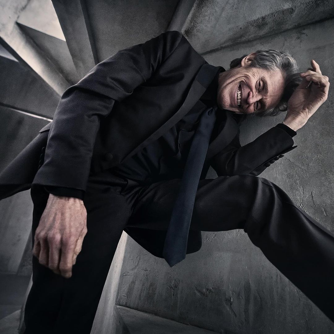 Middle-aged man in black suit and tie leans dramatically against a concrete spiral staircase with a happy facial expression.
