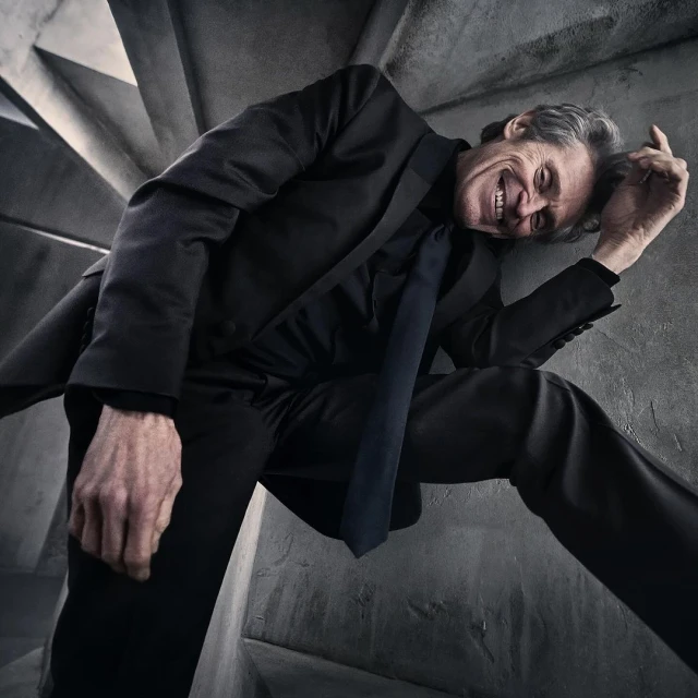 Middle-aged man in black suit and tie leans dramatically against a concrete spiral staircase with a happy facial expression. 