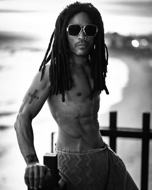 Black and white portrait of shirtless Lenny Kravitz with dreadlocks, sunglasses, and a towel, posing on a balcony with an ocean view.