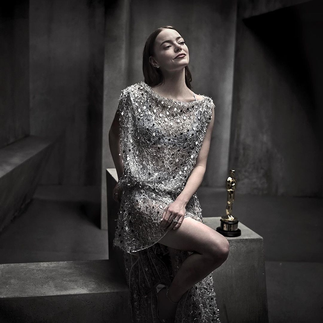 Adult female in an embellished dress poses in a photo studio with an Academy Award statue. Tonal grey background.