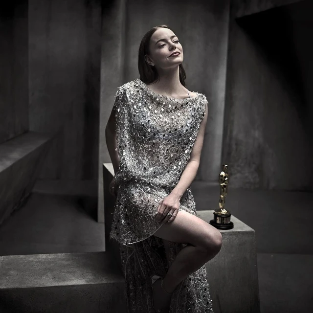 Adult female in an embellished dress poses in a photo studio with an Academy Award statue. Tonal grey background.
