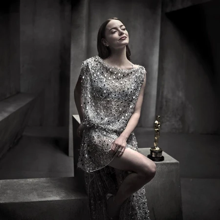Adult female in an embellished dress poses in a photo studio with an Academy Award statue. Tonal grey background.