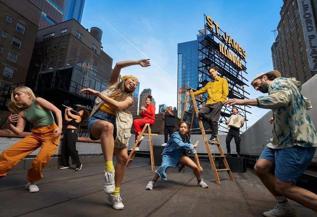 Group of eight people dancing on a rooftop in an urban setting, with city buildings, ladders, and musical instruments in the background.