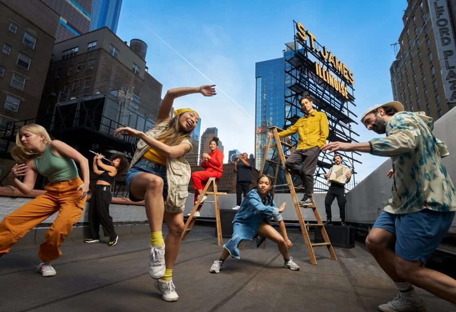 Group of eight people dancing on a rooftop in an urban setting, with city buildings, ladders, and musical instruments in the background.