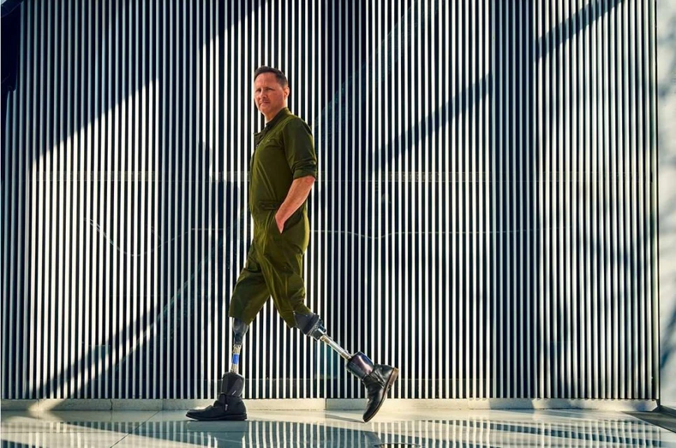 An adult male with prosthetic legs walks with his hand in his pocket in front of a wall with black and white vertical stripes.