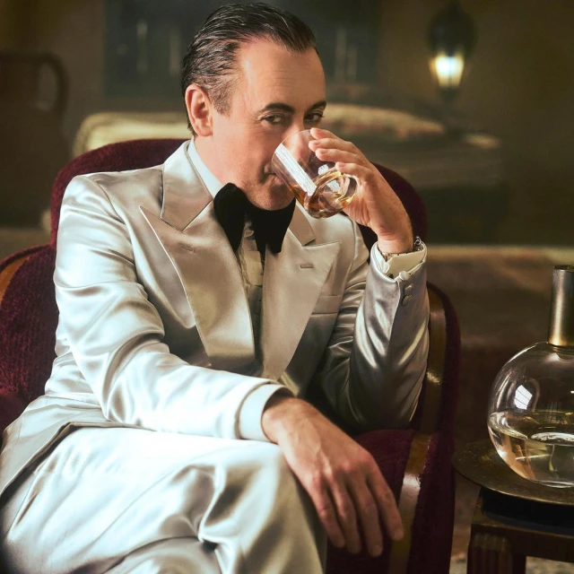 Elegant adult man in silver suit drinking whiskey indoors. Man seated in chair, holding glass, looking at the camera in moody lighting.