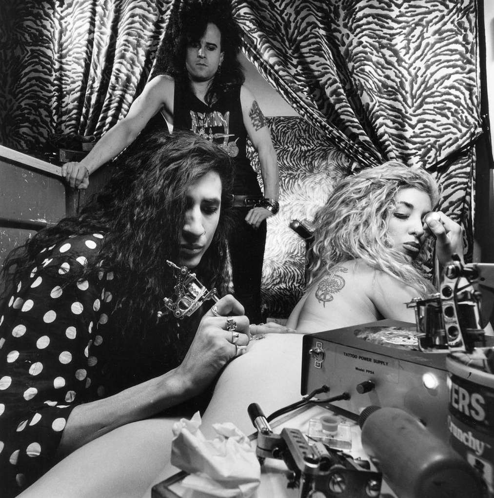 Black and white image of a tattoo artist working on a client's back, with another person standing by, in a room with patterned curtains.