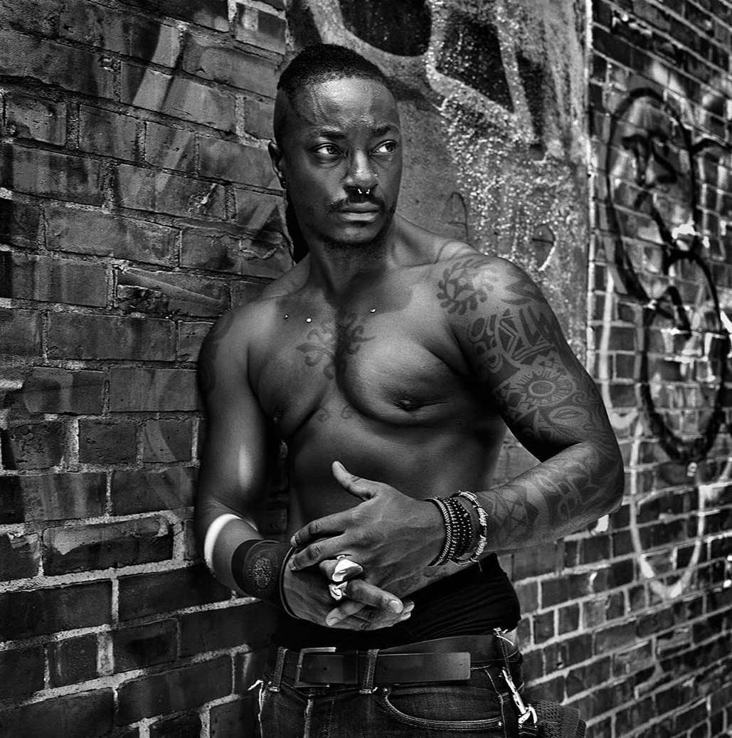 Black and white shot of a muscular, tattooed adult male with jewelry posing against a brick wall with graffiti in an urban setting.