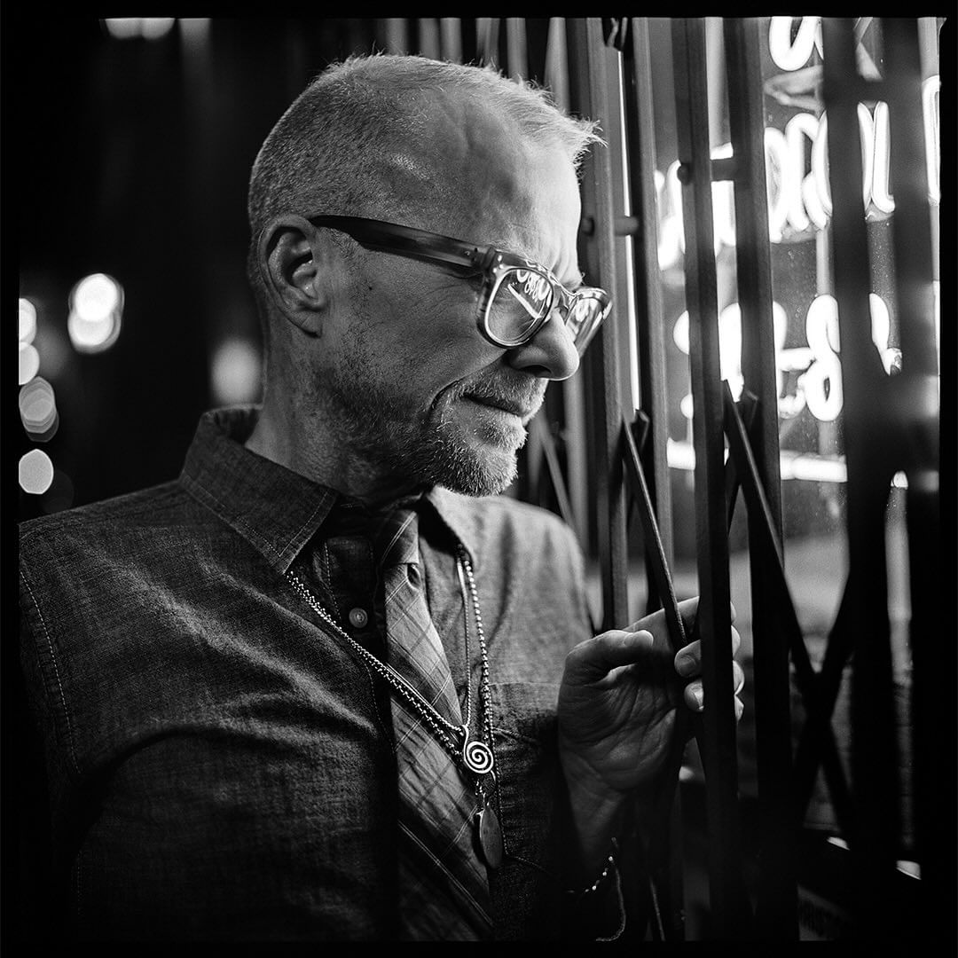 Black and white portrait of a man in glasses wearing a button-down shirt and necklaces, holding onto metal bars.
