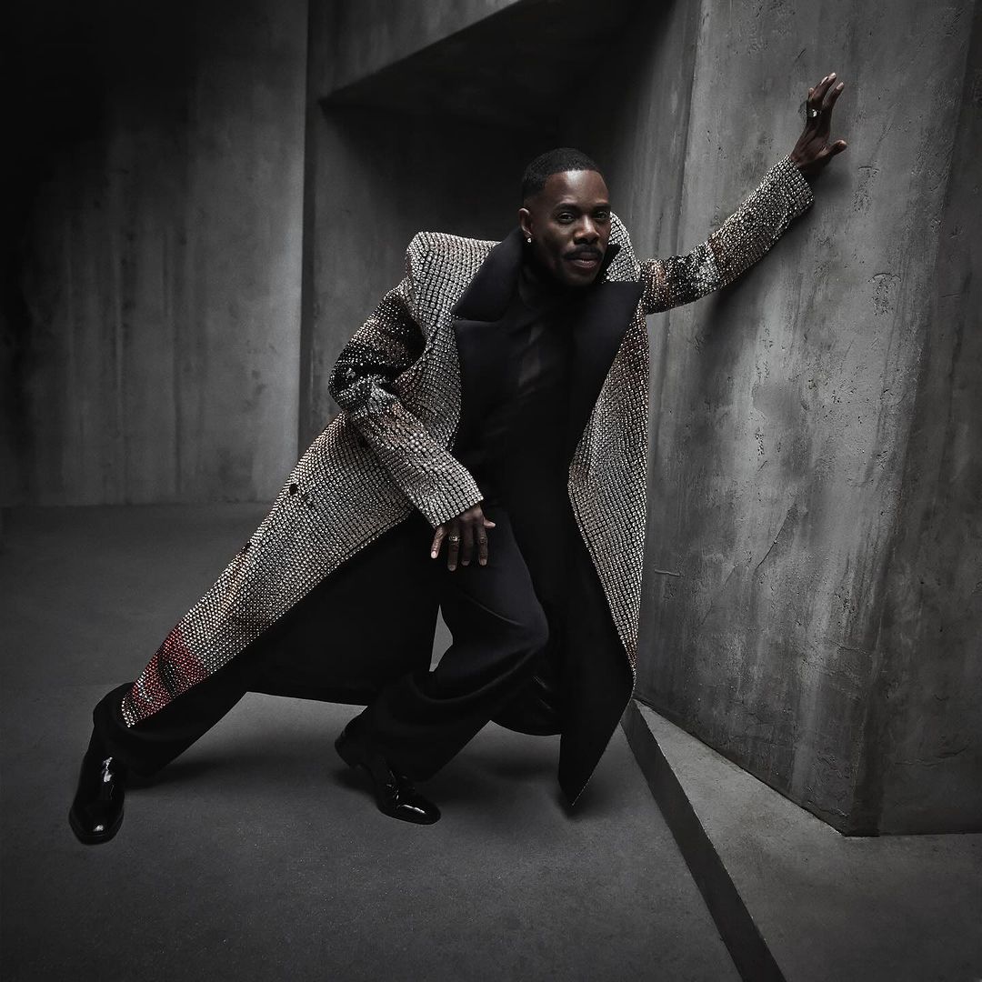 A dark-skinned man in a patterned overcoat leans against a concrete wall wearing black pants and a black turtle neck.