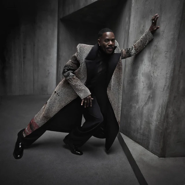 A dark-skinned man in a patterned overcoat leans against a concrete wall wearing black pants and a black turtle neck.