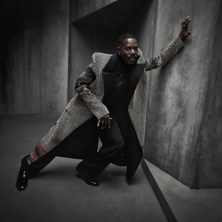 A dark-skinned man in a patterned overcoat leans against a concrete wall wearing black pants and a black turtle neck.