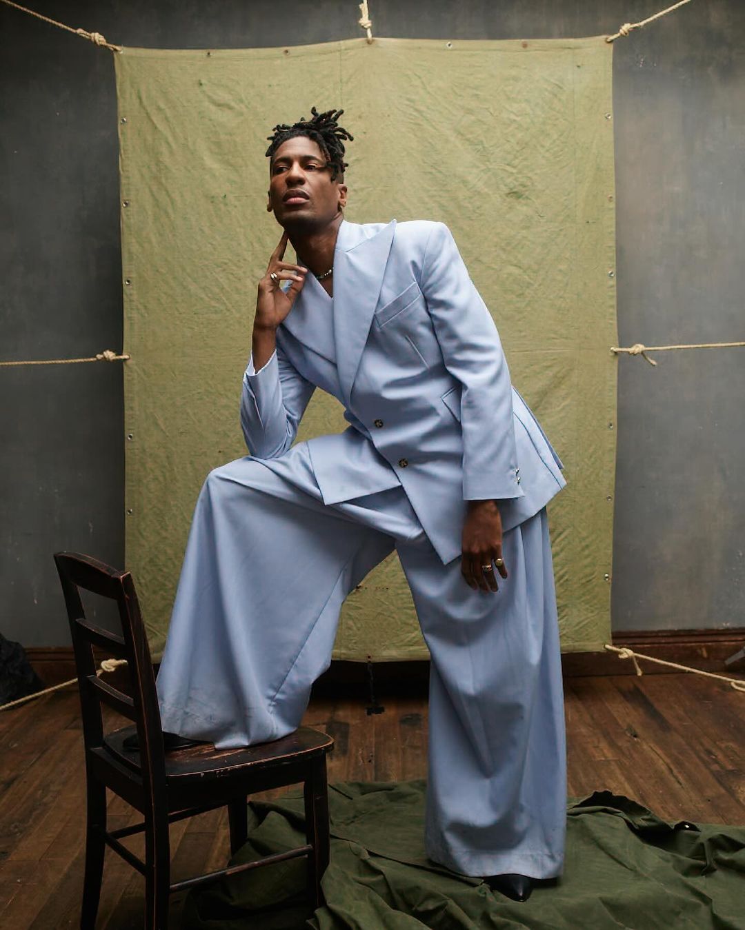 Man in a light blue double-breasted suit poses on a wooden chair against a textured backdrop in a fashion forward editorial.
