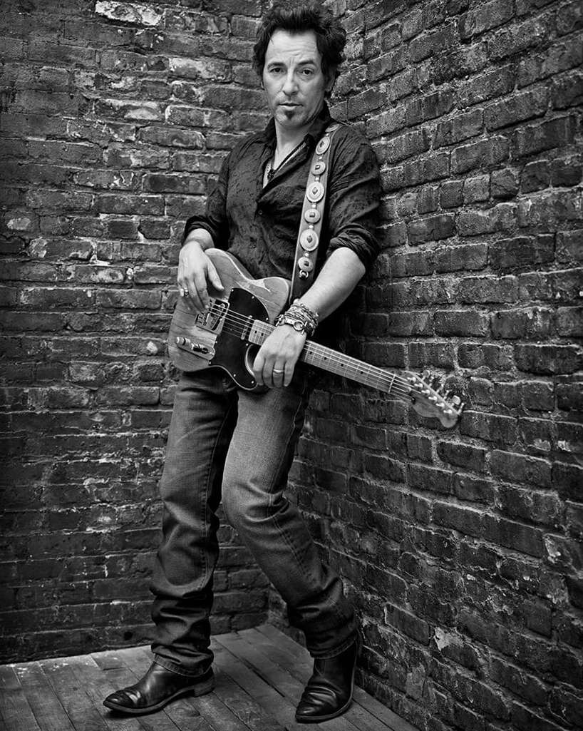 Black and white portrait of musician Bruce Springsteen standing against a brick wall, holding his guitar, looking at the camera.