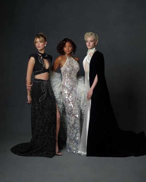 Three women in formal gowns pose in a studio. The woman on the left wears a black crop top and skirt, the woman in the center, a silver gown. The woman on the right, a black dress.
