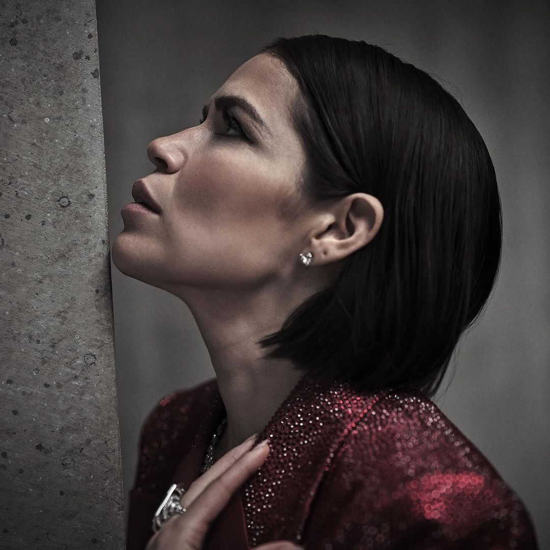 Adult woman with short dark hair poses in a red sequined jacket while leaning against a concrete wall in a close up shot.