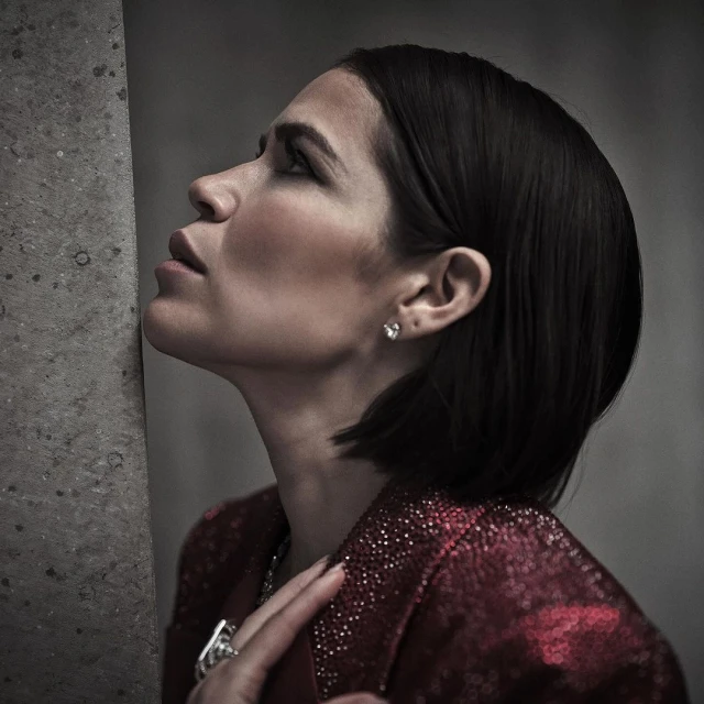 Adult woman with short dark hair poses in a red sequined jacket while leaning against a concrete wall in a close up shot.