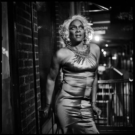 Black woman wearing a sleeveless evening gown and blonde wig posing outdoors at night against a brick wall, photographed by Mark Seliger