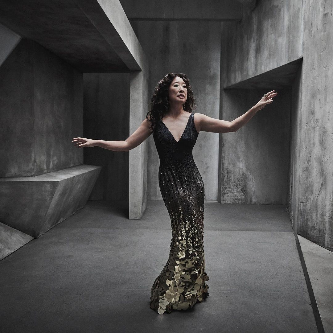 Sandra Oh strikes an elegant pose in a black and gold sequined gown against a geometric, concrete backdrop, adding a touch of sophistication.