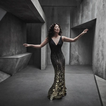 Sandra Oh strikes an elegant pose in a black and gold sequined gown against a geometric, concrete backdrop, adding a touch of sophistication.