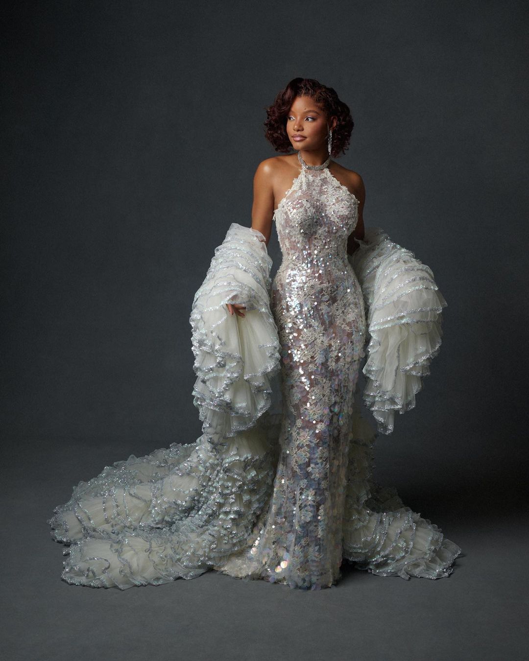 Woman poses in a silver halter gown with white ruffled sleeves and a matching floor-length train against a grey studio backdrop.