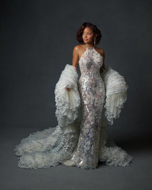 Woman poses in a silver halter gown with white ruffled sleeves and a matching floor-length train against a grey studio backdrop.