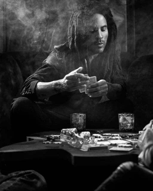 Monochrome portrait of a Black adult male with dreadlocks, playing cards at a poker table surrounded by chips and whiskey glasses, indoors.