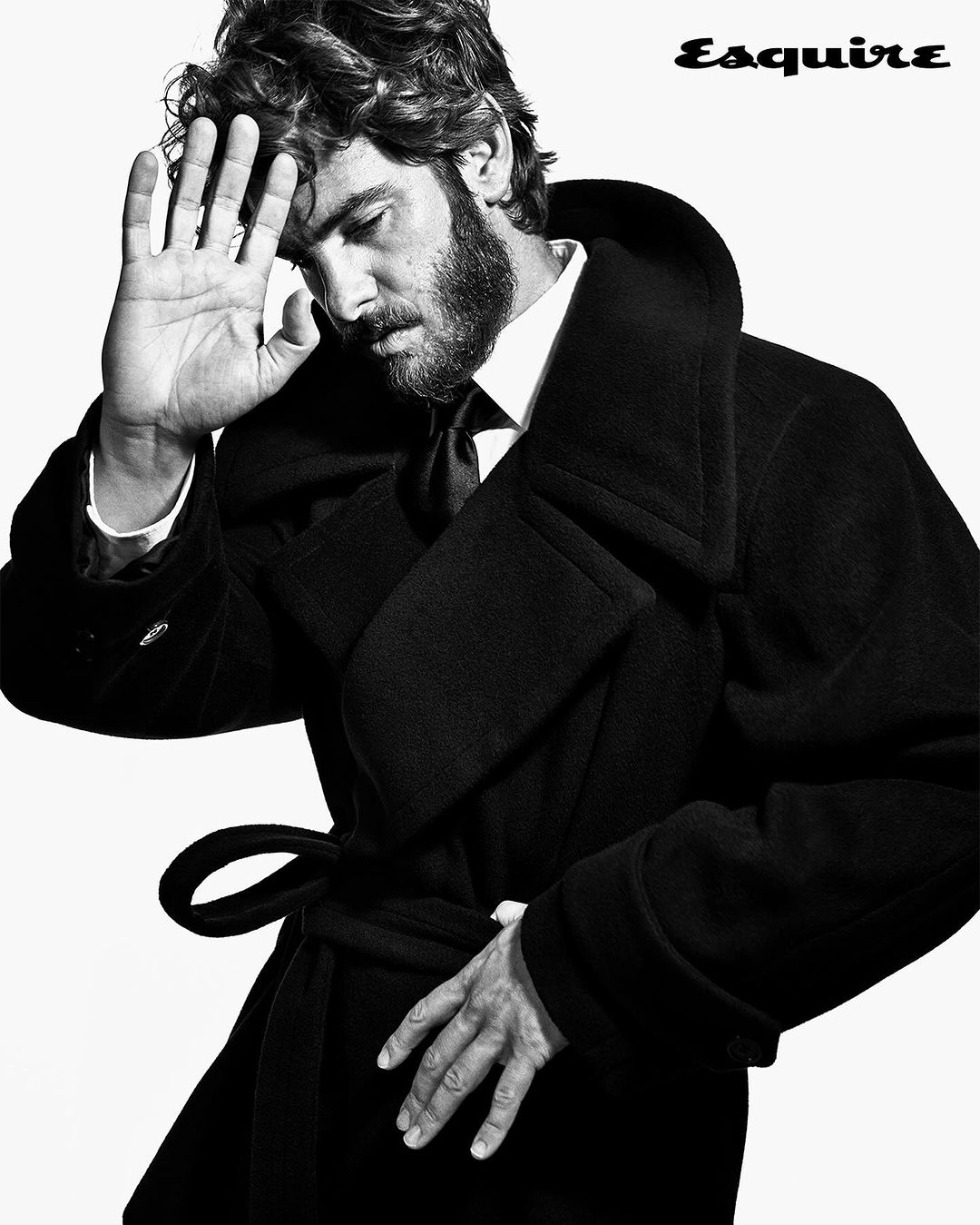 Black-and-white portrait of a man in a wool coat looking down with his hand raised to his forehead, Esquire magazine logo.