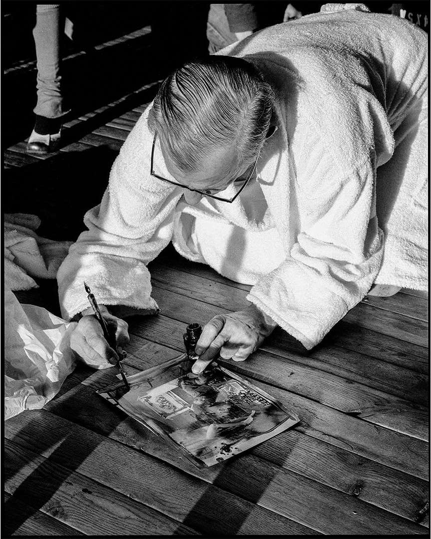 Black and white shot of an adult leaning over a magazine with a pen and ink bottle on a wood plank surface in an indoor environment.