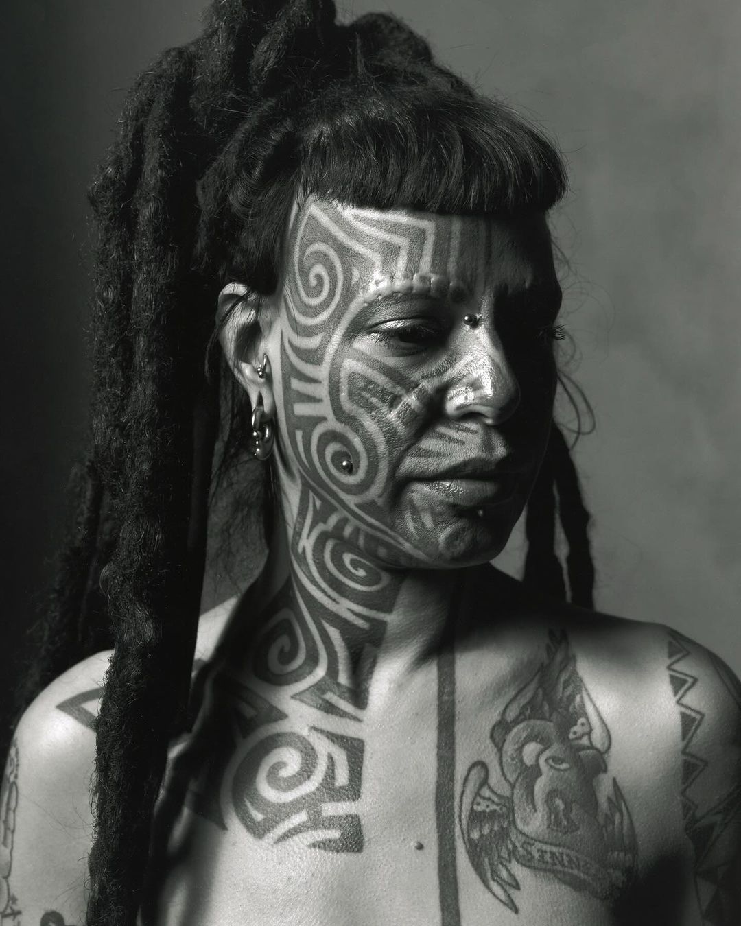 Striking black and white portrait of a woman with dreadlocks, distinctive face tattoos, and multiple piercings, creating a unique aesthetic.