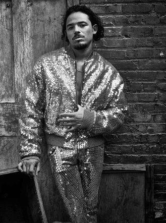 Monochrome portrait of a man in a sequined jacket and pants leaning against a wall, showcasing fashion, style, and urban ambiance.