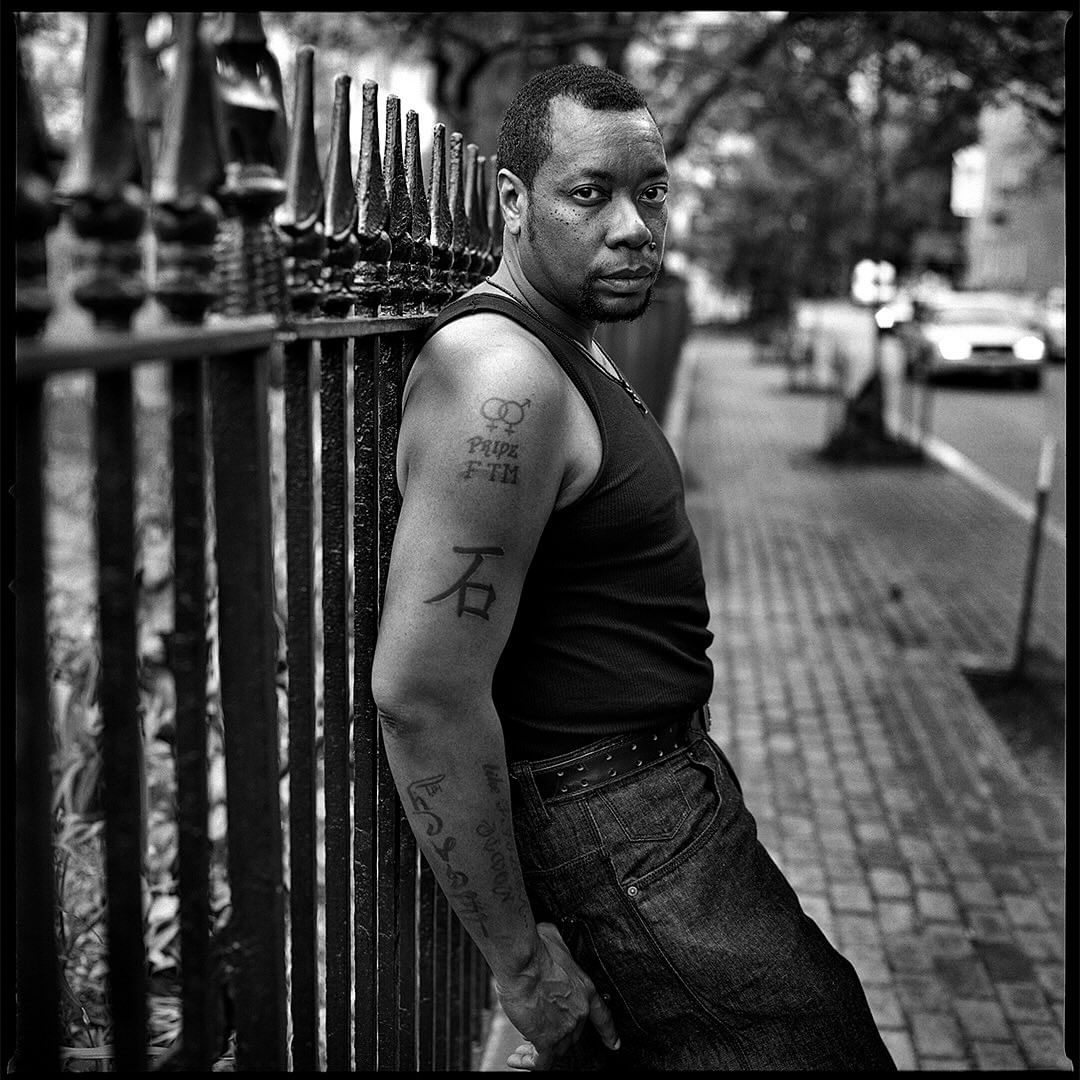 Portrait of a man with tattoos leaning on a wrought iron fence in a black and white photograph with trees and buildings in the background.