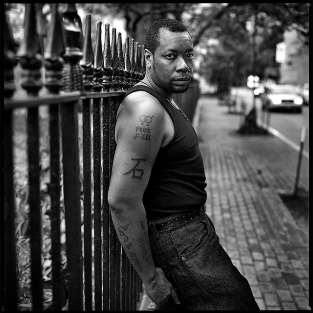 Portrait of a man with tattoos leaning on a wrought iron fence in a black and white photograph with trees and buildings in the background.