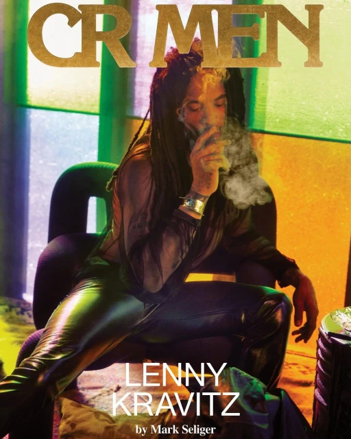 Lenny Kravitz sits in a chair, smoking with long dreadlocks and a sheer top, against an orange and green background on magazine cover.