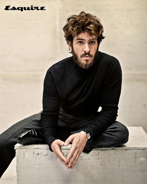 Portrait of Andrew Garfield, seated on a concrete block, hands clasped, wearing a black textured sweater and gray slacks, shot in studio.