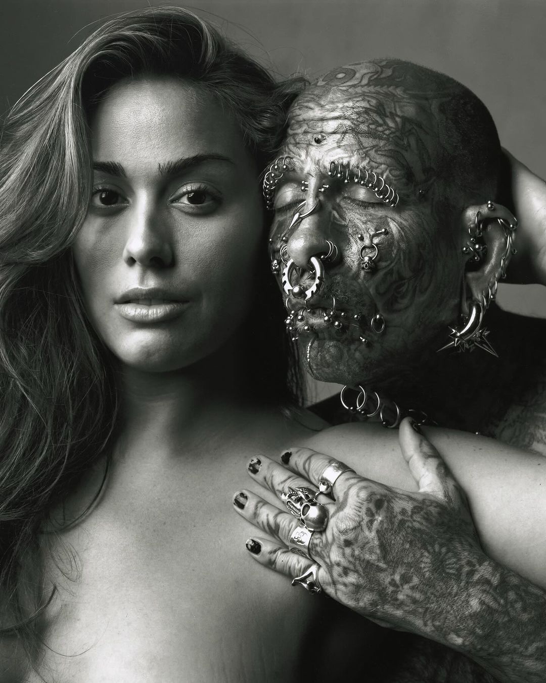Monochrome portrait of a young woman with long wavy hair standing next to a man with full-body tattoos and facial piercings in a studio setting.