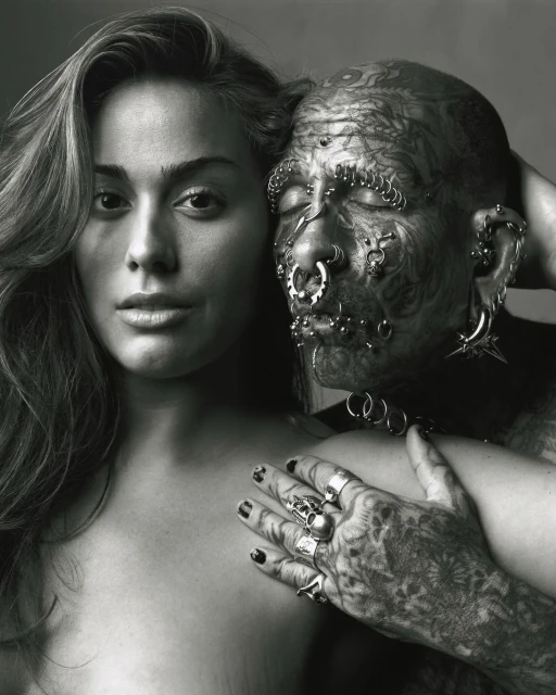 Monochrome portrait of a young woman with long wavy hair standing next to a man with full-body tattoos and facial piercings in a studio setting.