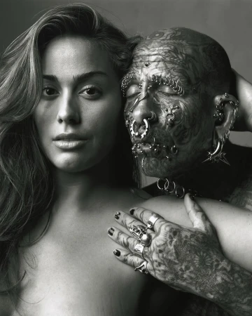 Monochrome portrait of a young woman with long wavy hair standing next to a man with full-body tattoos and facial piercings in a studio setting.