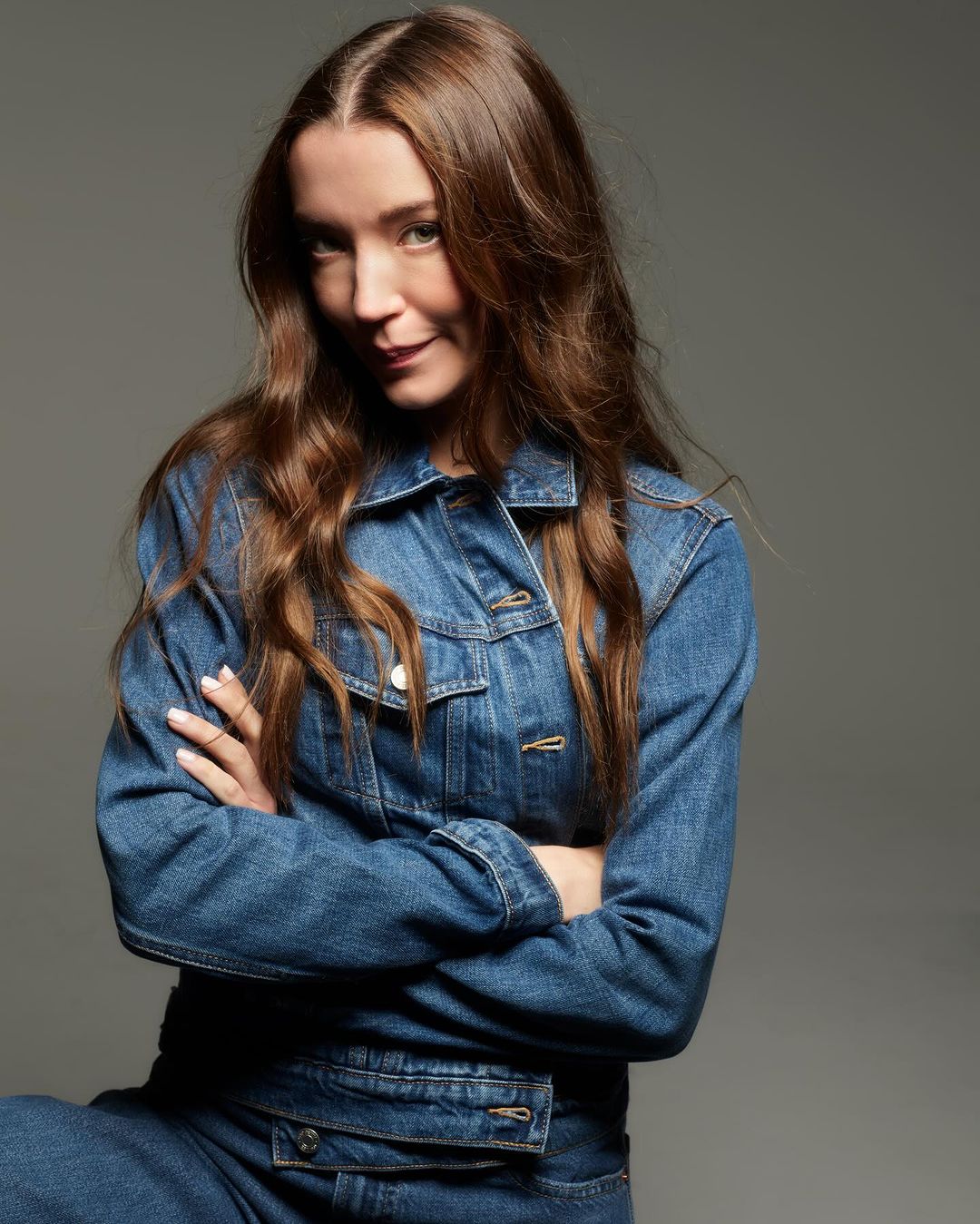 Woman in denim jacket with arms crossed posing against a grey seamless studio background