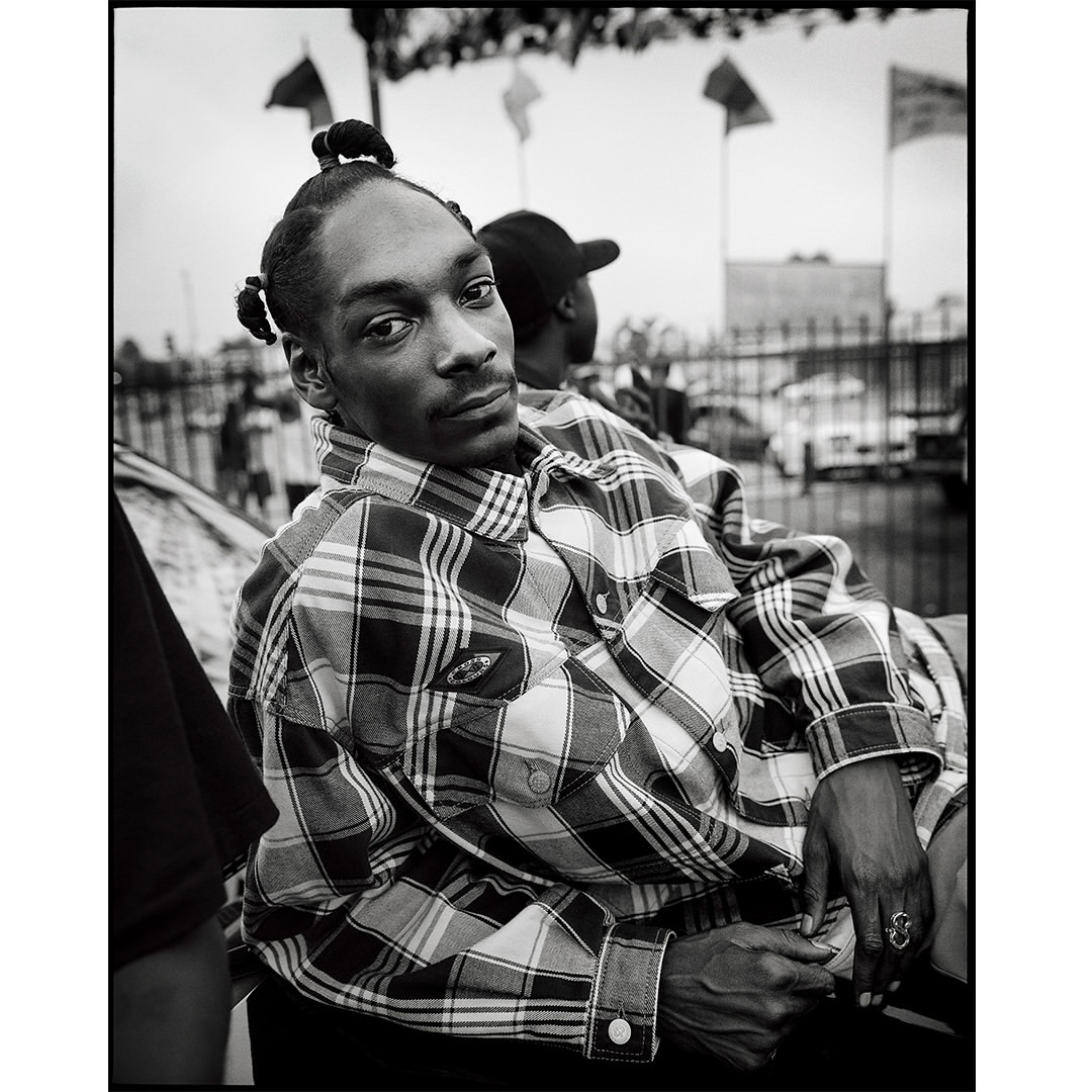 Monochrome portrait of Snoop Dogg in a plaid shirt, seated outdoors with a second person behind him and flags in the background.