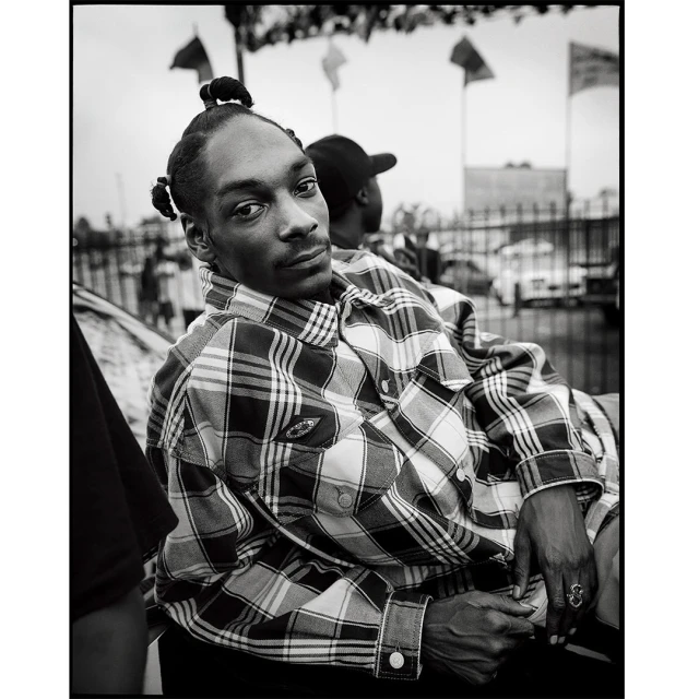 Monochrome portrait of Snoop Dogg in a plaid shirt, seated outdoors with a second person behind him and flags in the background.