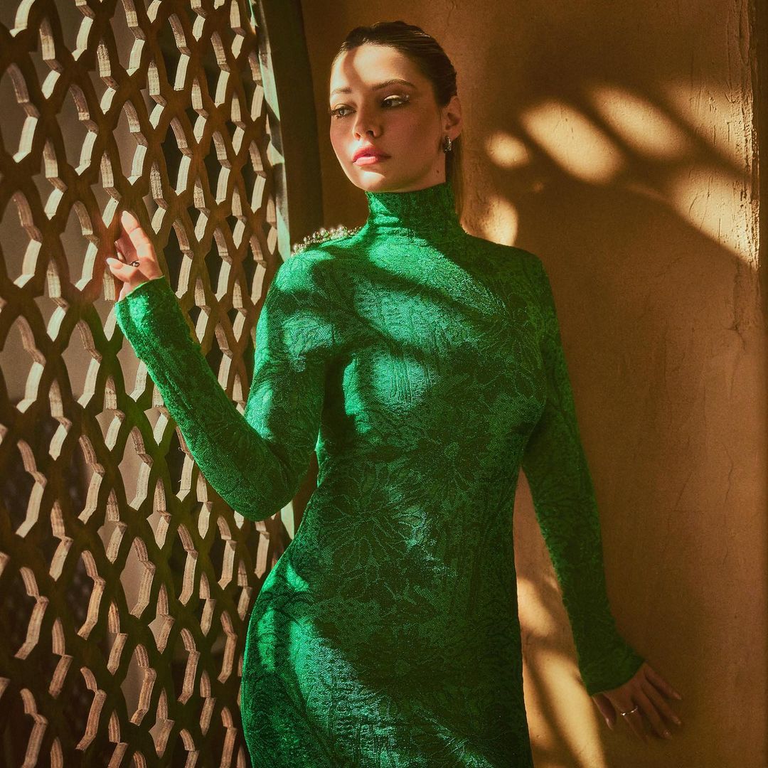 Woman in emerald green dress posing near lattice screen. Portrait shot with warm lighting and shadow play on textured wall.