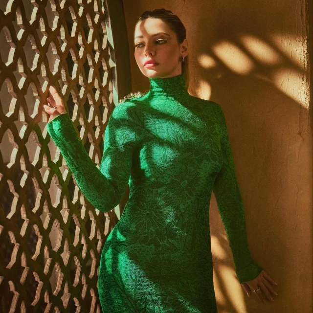 Woman in emerald green dress posing near lattice screen. Portrait shot with warm lighting and shadow play on textured wall.
