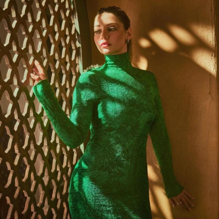 Woman in emerald green dress posing near lattice screen. Portrait shot with warm lighting and shadow play on textured wall.