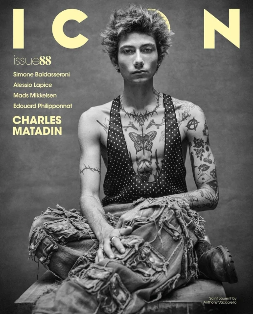 Magazine cover featuring a young adult male with tattoos, wearing a vest and pants, posed in a studio against a grey background.